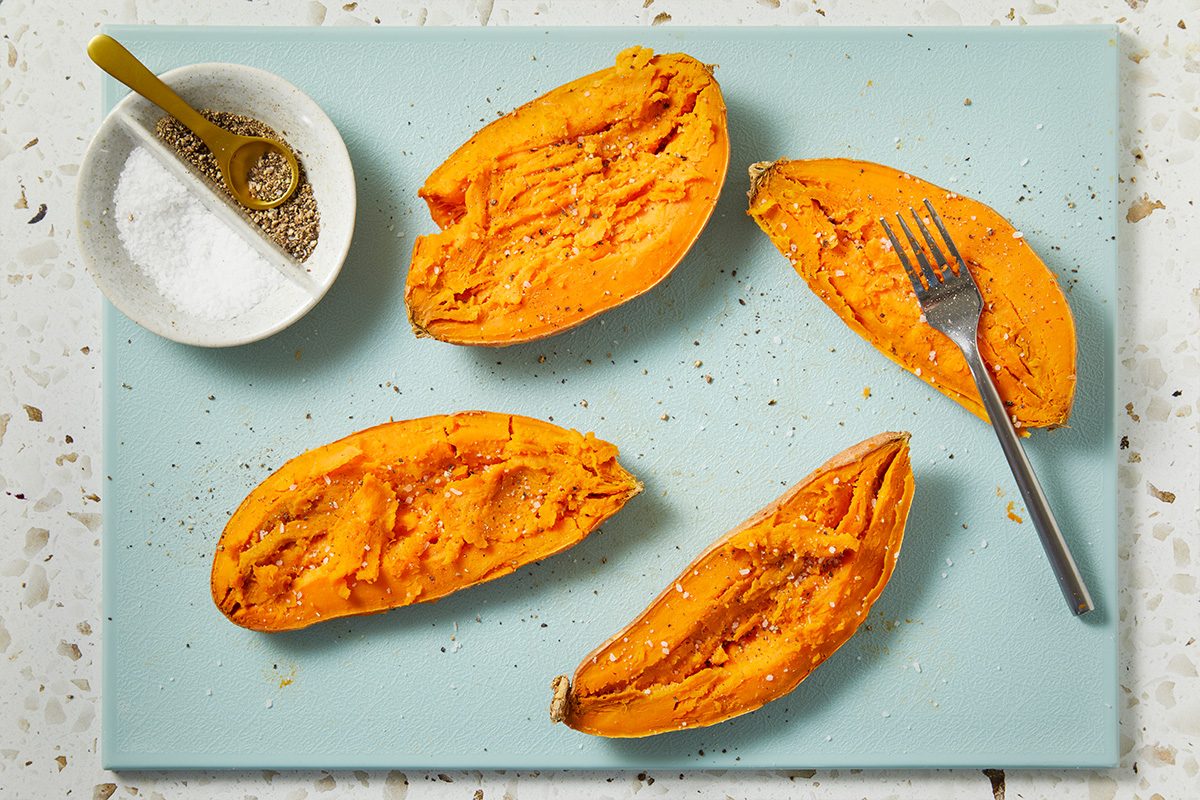 Horizontal overhead image showing sweet potatoes split open and fluffed, revealing their vibrant orange interior. A spoon rests nearby, suggesting preparation for seasoning and smashing.