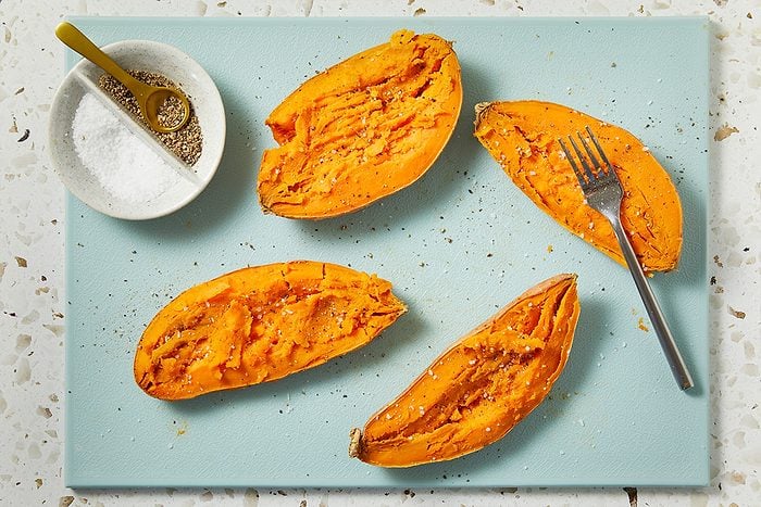Horizontal overhead image showing sweet potatoes split open and fluffed, revealing their vibrant orange interior. A spoon rests nearby, suggesting preparation for seasoning and smashing.