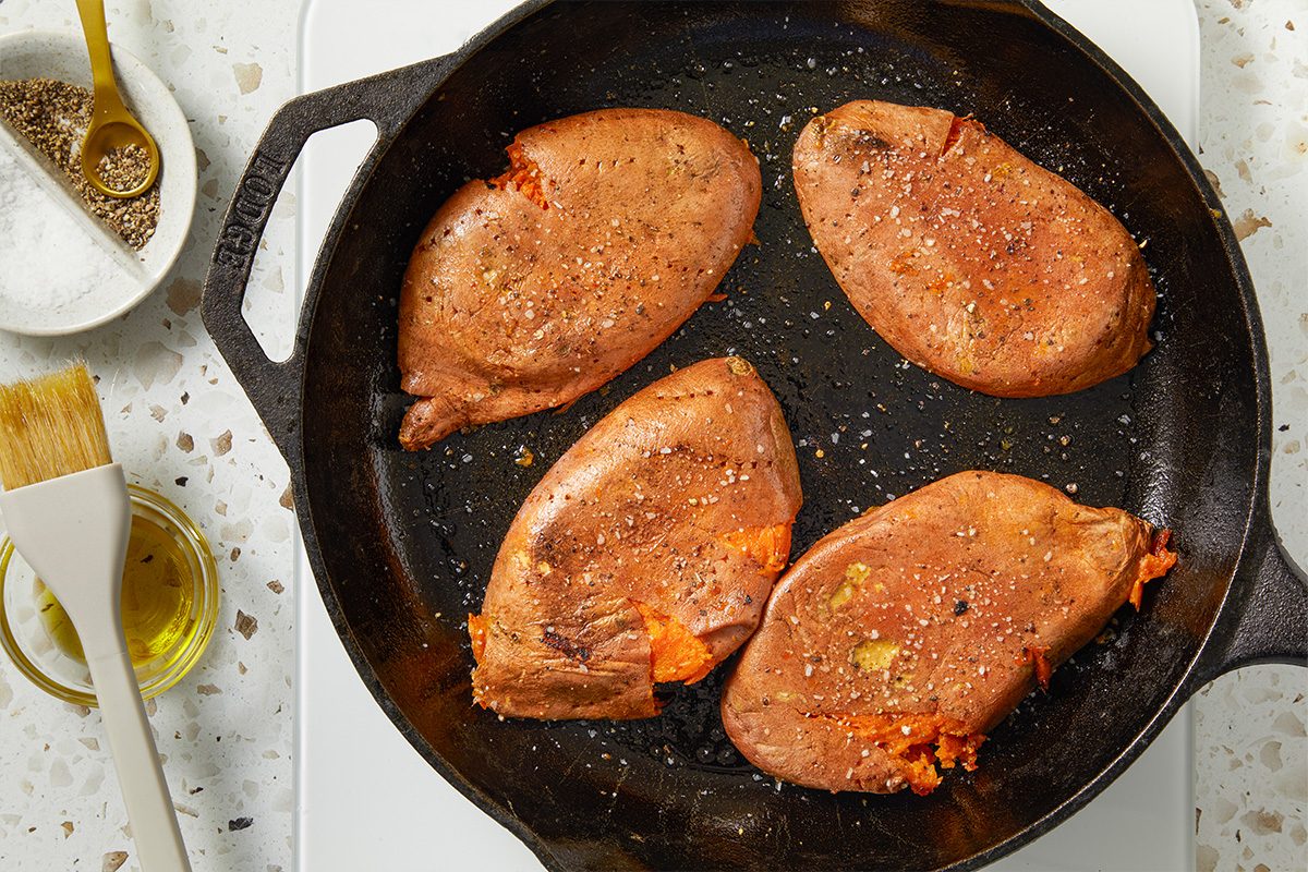 Overhead horizontal image of sweet potato halves placed cut-side down in a cast-iron skillet. The composition shows the beginning of the browning process before topping with cheese and herbs.