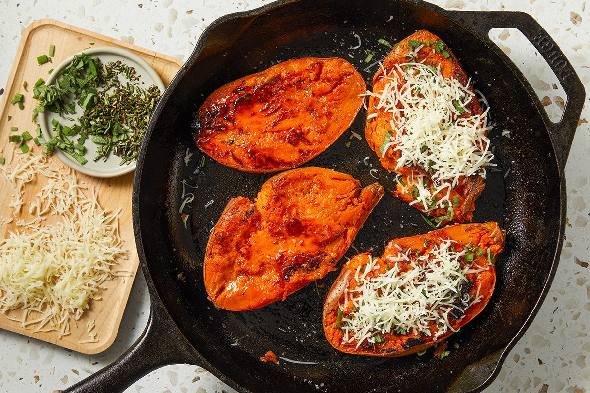 Horizontal overhead image of smashed sweet potatoes in a skillet topped with finely grated Gruyere cheese and fresh herbs. The cheese is beginning to melt, signaling the transition to the final bake.