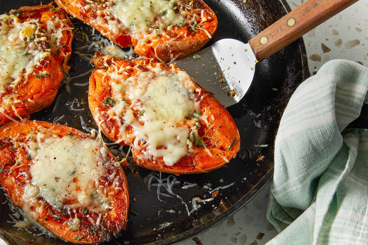 Horizontal close-up of smashed sweet potatoes in a skillet as a spatula lifts one portion. Melted Gruyere stretches slightly, highlighting texture and crisp edges.