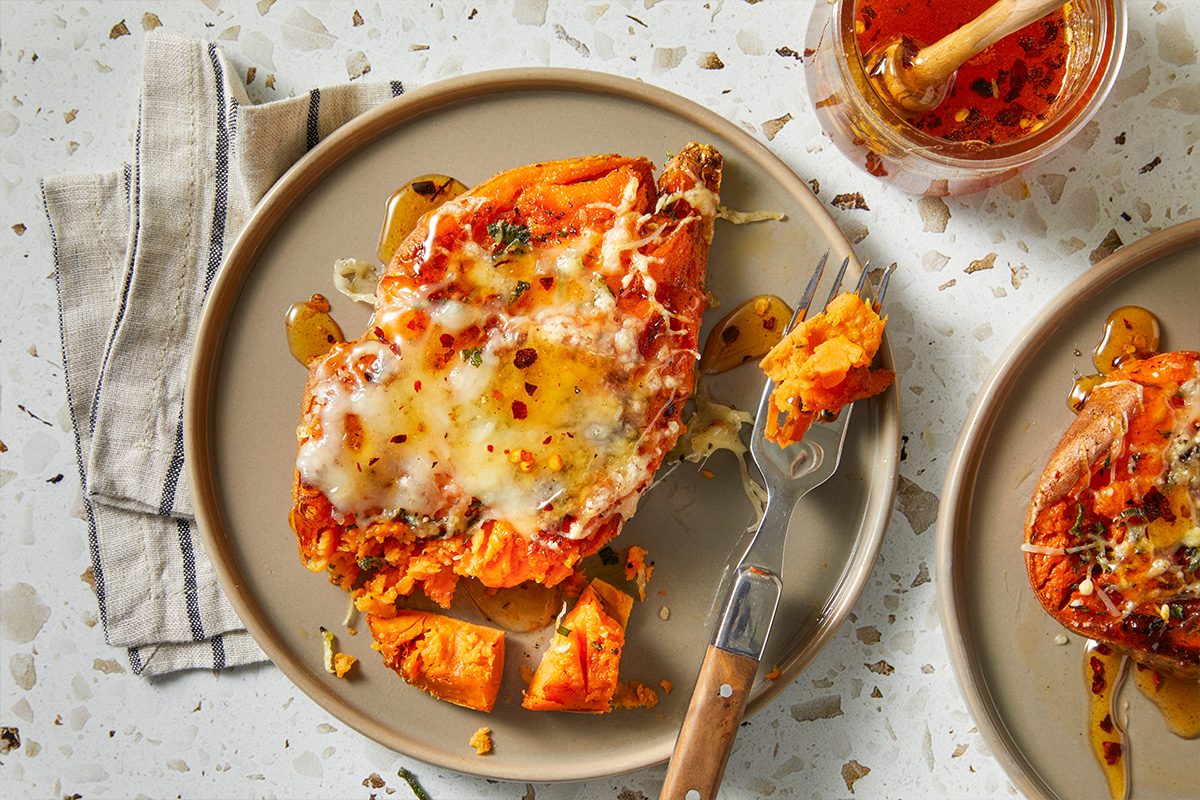 Horizontal overhead serving shot of a plated smashed sweet potato topped with melted Gruyere, herbs and a drizzle of hot honey. A fork rests nearby, showcasing the soft interior and crisp exterior contrast.