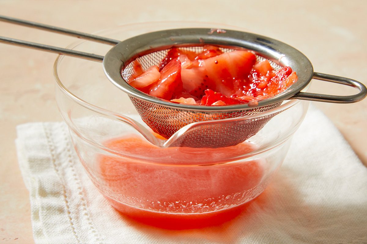 Overhead horizontal image of strained strawberry puree passing through a sieve into a bowl for cocktail mixing.