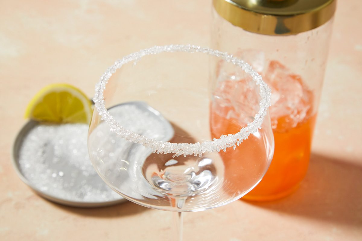 3/4 angle horizontal image of a coupe glass being rimmed with sugar for a strawberry lemon drop cocktail.