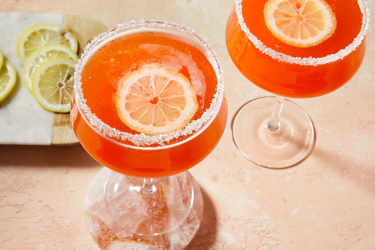 Eye-level horizontal image of two strawberry lemon drop cocktails served in coupe glasses with sugared rims and lemon garnish.