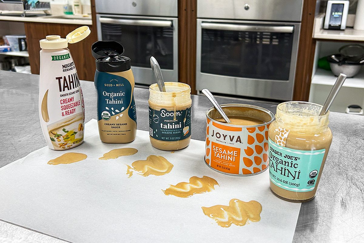Five different tahini brands, each with a spoon, are arranged on a counter. Each one has a small puddle of tahini in front of it on white paper, showcasing differences in color and texture. Kitchen appliances are visible in the background.