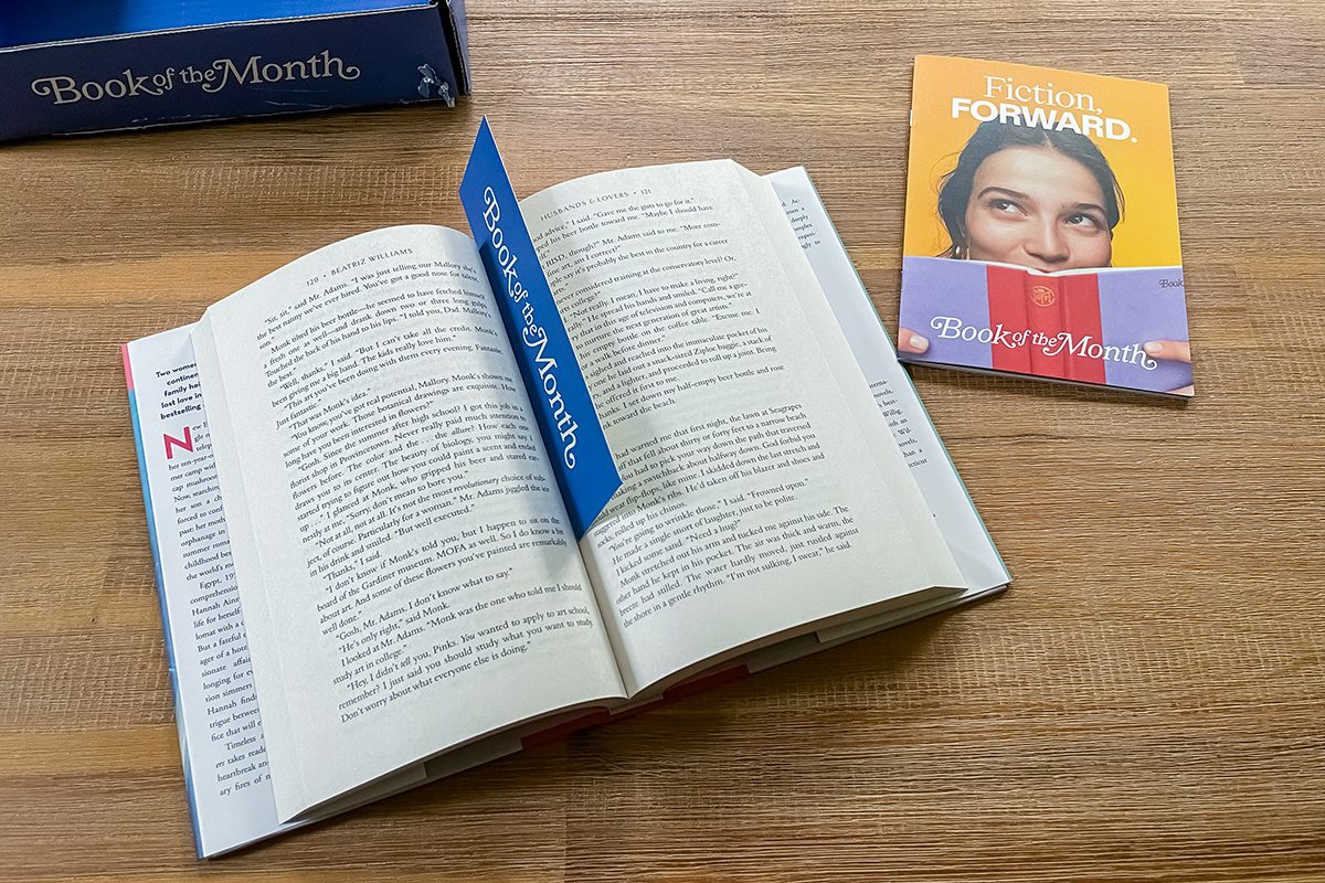 An open book with a "Book of the Month" bookmark lies on a wooden table. Nearby is a yellow booklet with a woman