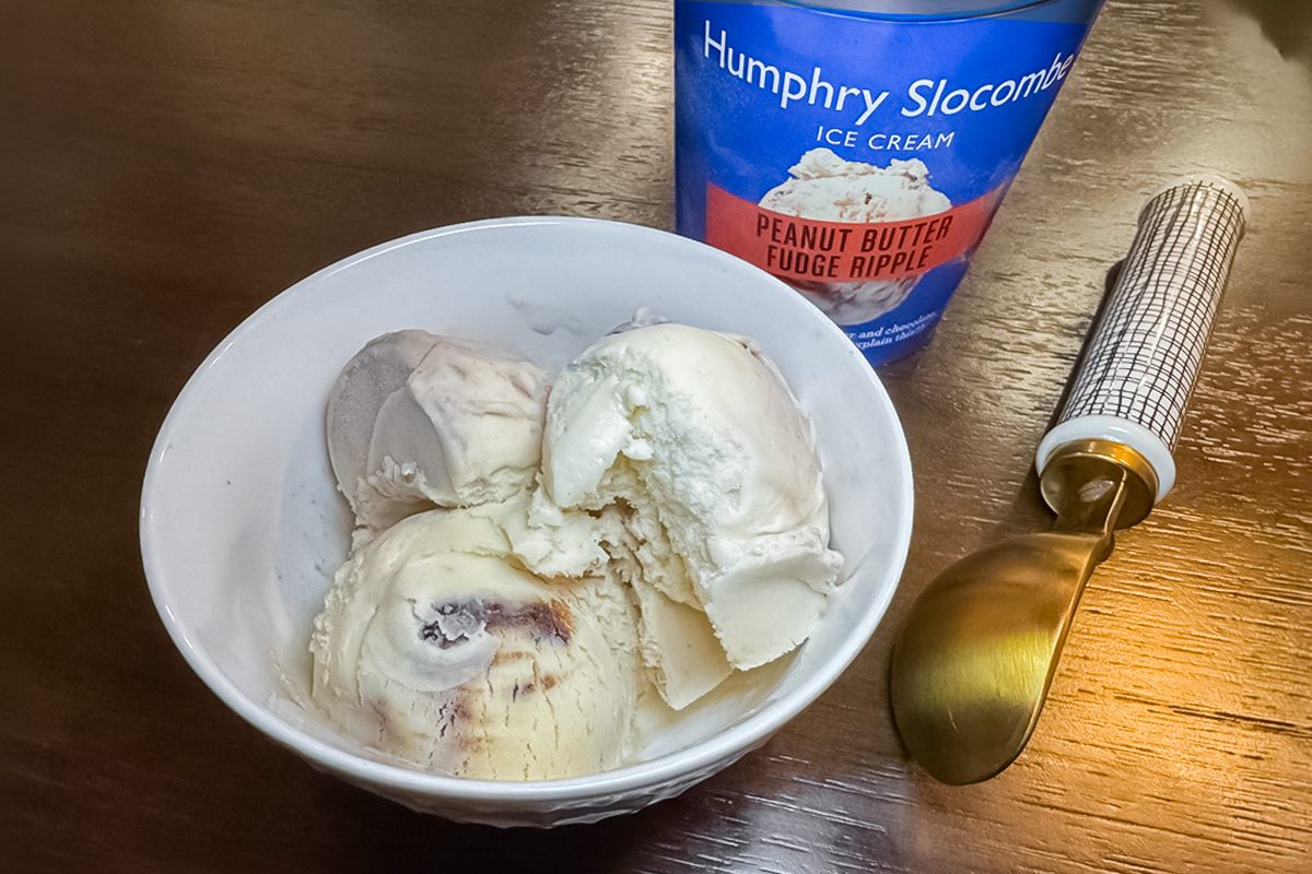 A white bowl with scoops of ice cream sits on a wooden table next to a gold ice cream scooper and a container labeled "Humphry Slocombe Ice Cream Peanut Butter Fudge Ripple.