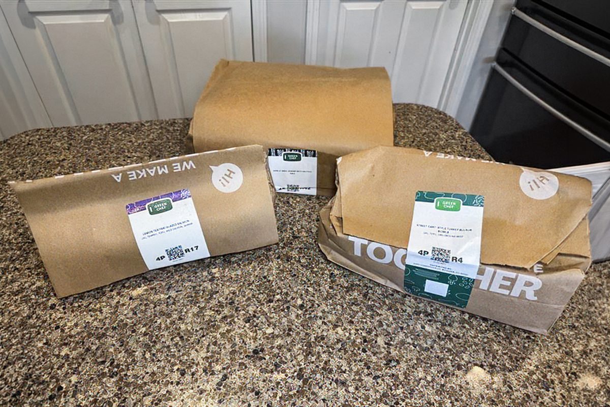Three brown paper-wrapped meal kit packages with labels are on a granite kitchen counter, with white cabinets and a stainless steel oven in the background.