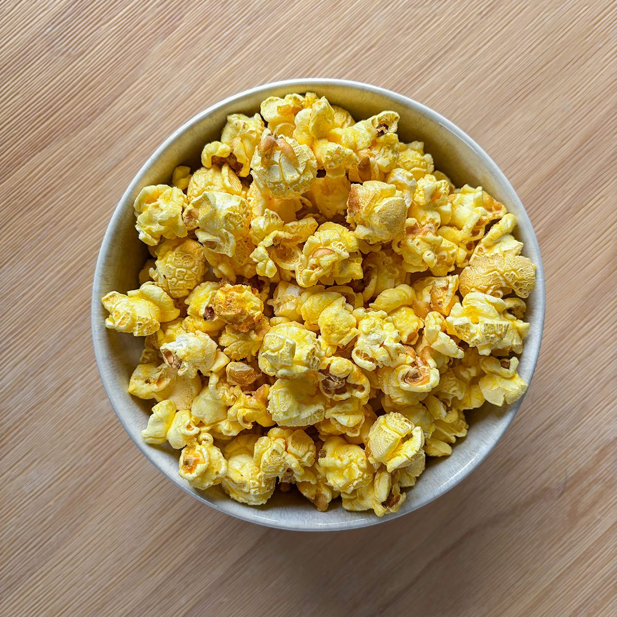 A white bowl filled with yellow, buttery popcorn sits on a light wooden surface.