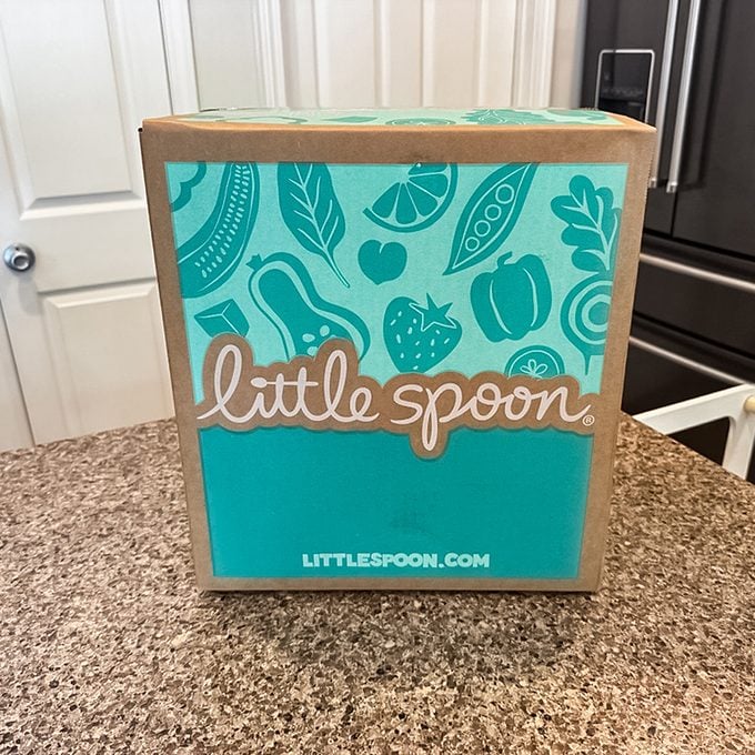 A Little Spoon delivery box with a teal and tan design sits on a kitchen counter. The box displays the brand name "Little Spoon" and illustrations of fruits and vegetables.