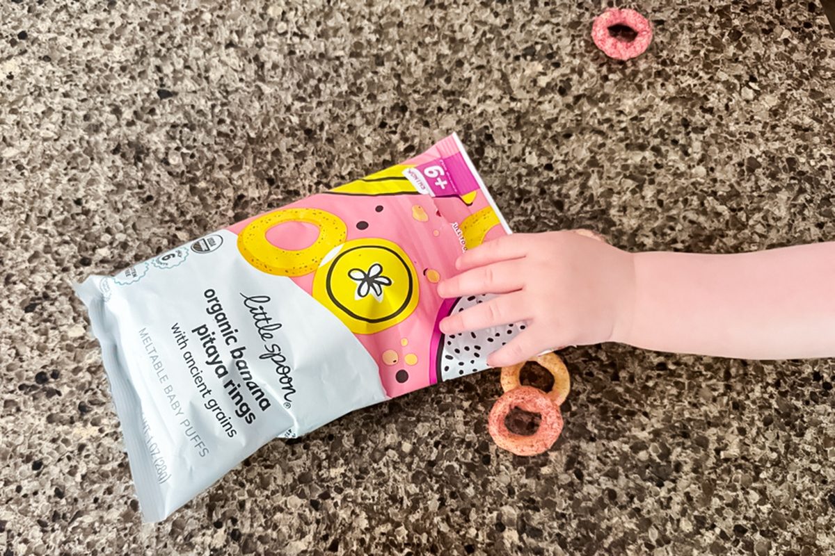 A child's hand reaches for a bag of Little Spoon organic beet berry finger foods on a speckled countertop, with a few round snacks scattered nearby.