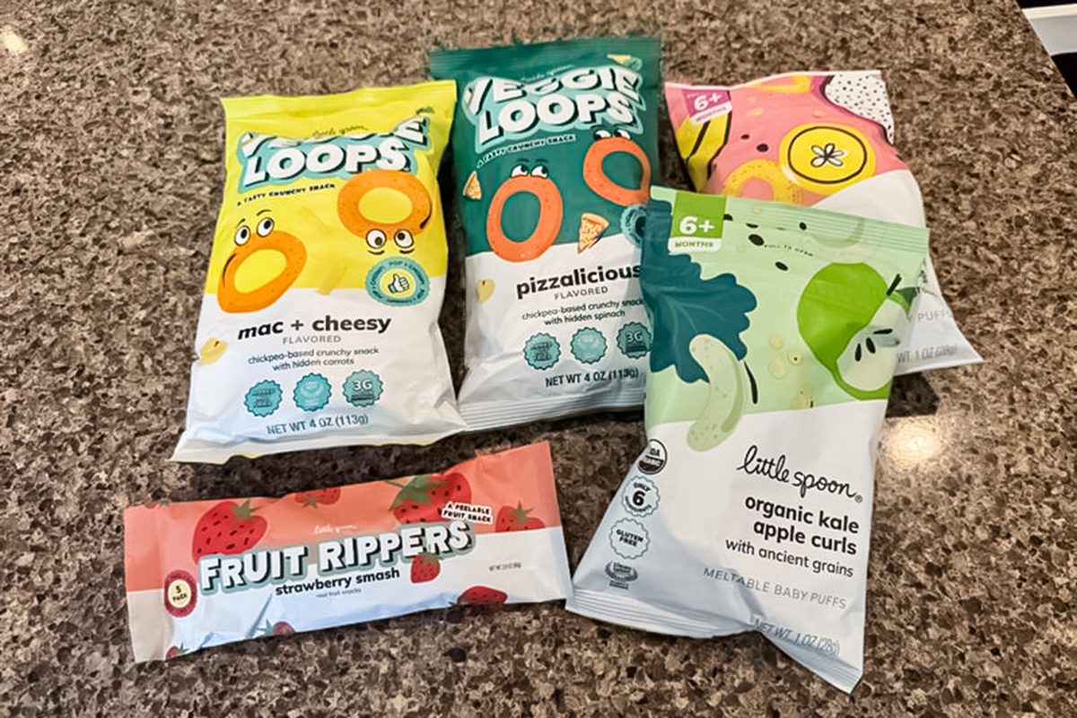 Five colorful snack packages on a granite countertop, including two bags of Little Spoon Loops (mac & cheesy and pizzalicious), two bags of organic kale apple curls, and one Fruit Rippers strawberry snack.