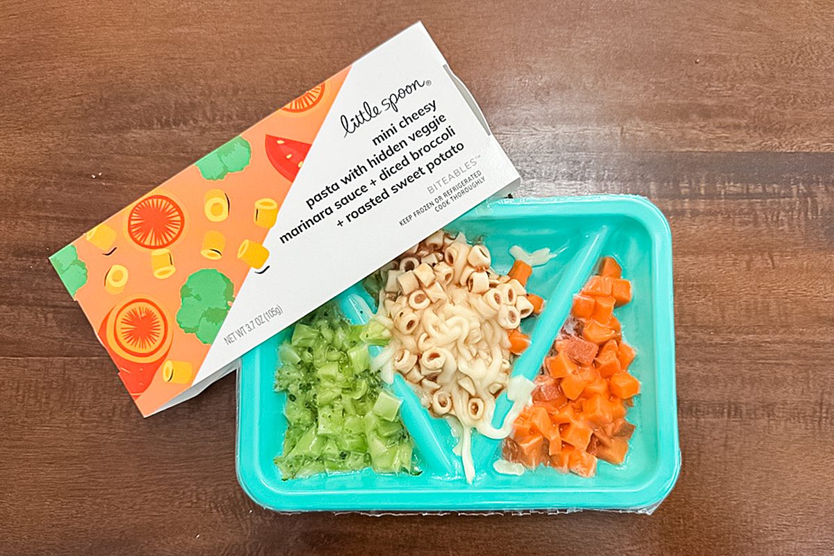 A divided turquoise tray holds chopped green vegetables, pasta with sauce, and diced orange vegetables. Above it is a carton labeled "Little Spoon" with an illustrated fruit and veggie design. Both are on a wooden surface.