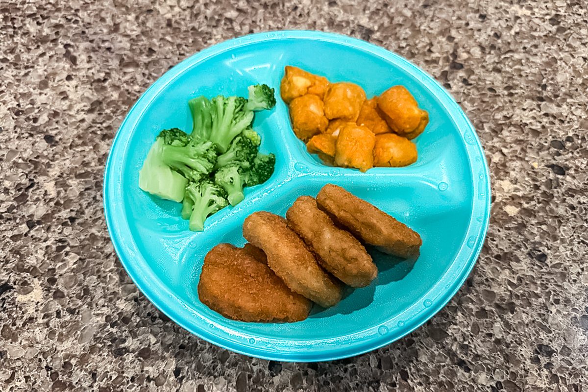 A blue divided plate with broccoli, orange macaroni and cheese, and three chicken nuggets, placed on a speckled gray countertop.