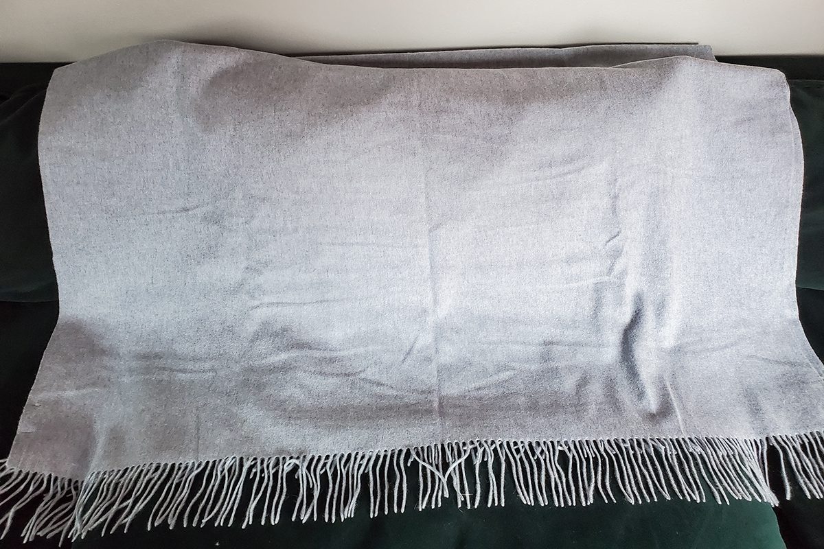 A light gray blanket with fringed edges is spread out on a dark-colored surface, possibly a couch or chair.
