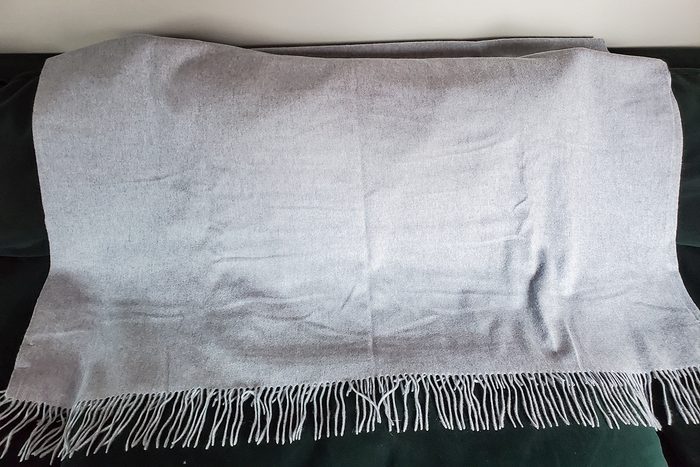 A light gray blanket with fringed edges is spread out on a dark-colored surface, possibly a couch or chair.