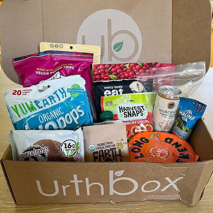 A cardboard UrthBox filled with assorted snacks, including veggie chips, popcorn, fruit crisps, a canned drink, a protein bar, and various healthy packaged foods, all neatly arranged inside the box.