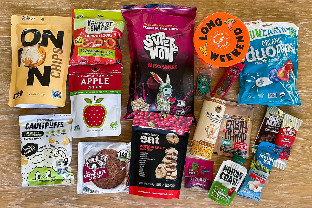A variety of colorful packaged snack foods, including chips, dried fruit, cookies, snack bars, puffs, and drink mixes, are spread out on a wooden surface. Each package is distinctly labeled and vibrant.