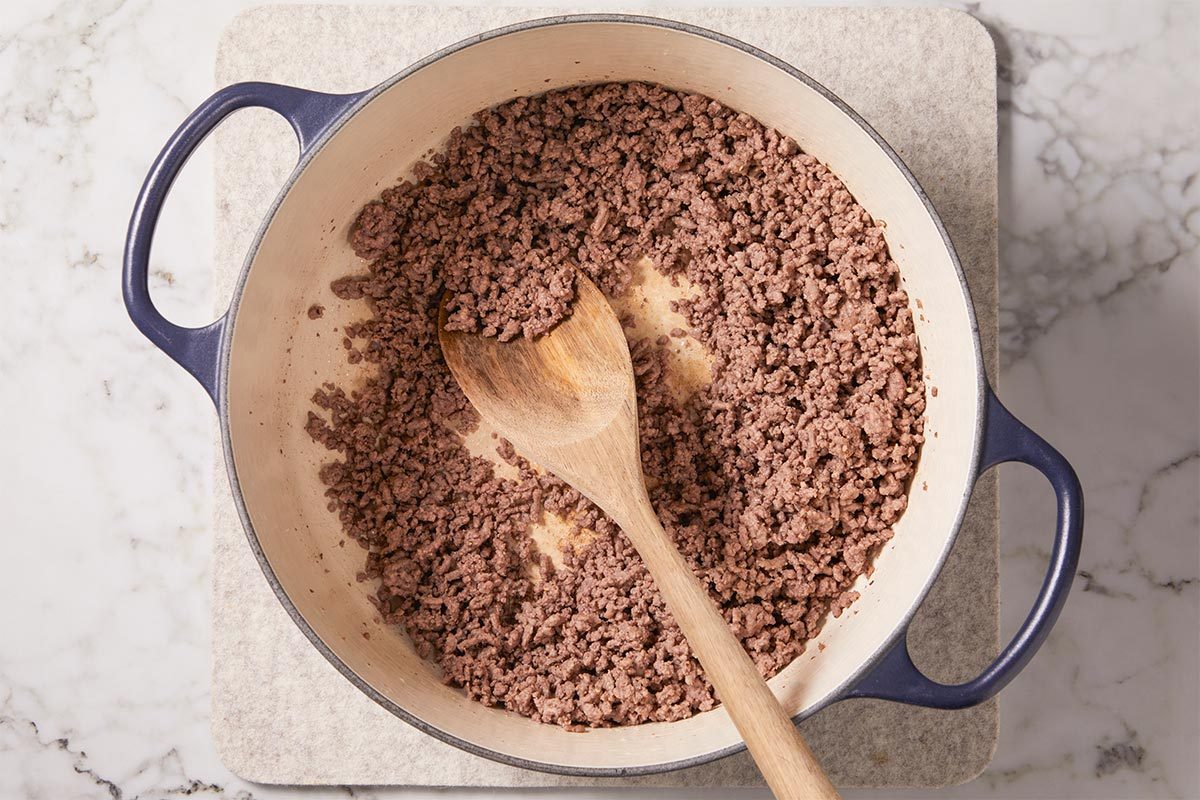 Overhead view of ground beef browning in a Dutch oven with a wooden spoon, beginning the base for Tagliatelle Bolognese. The beef is seasoned with salt and pepper and cooked until crumbly and no longer pink, forming the hearty foundation of this classic Italian pasta sauce.