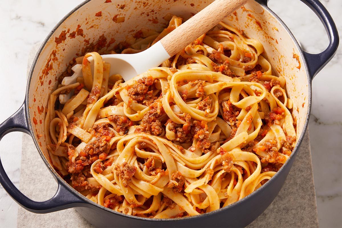 Overhead close-up of Tagliatelle Bolognese in a Dutch oven with pasta fully coated in rich meat sauce. The thick ragu clings to the wide noodles, showcasing the finished texture and consistency.