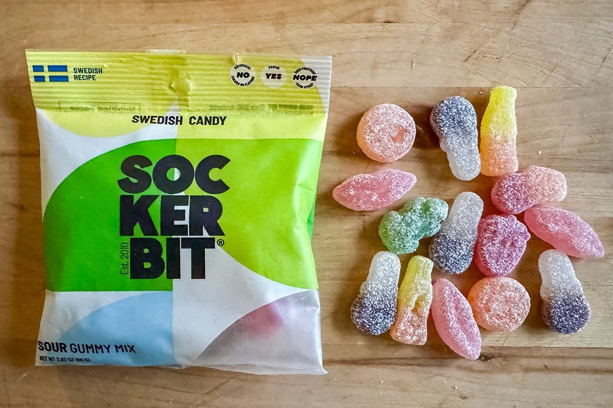 A bag of Swedish candy labeled “Sockerbit Sour Gummy Mix” sits on a wooden surface with assorted colorful gummy candies poured out next to it.