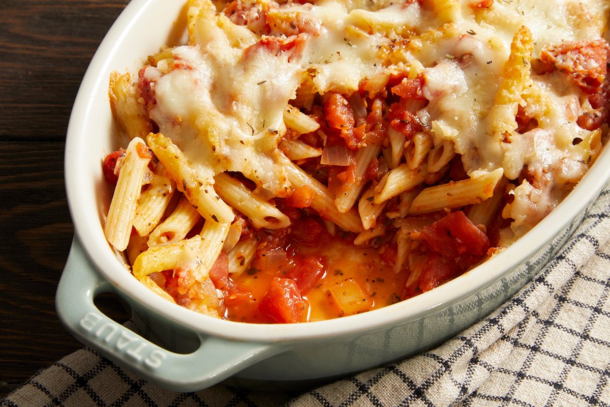 Baked penne pasta in a white casserole dish, topped with melted cheese and tomato sauce, sitting on a checkered cloth on a wooden surface.