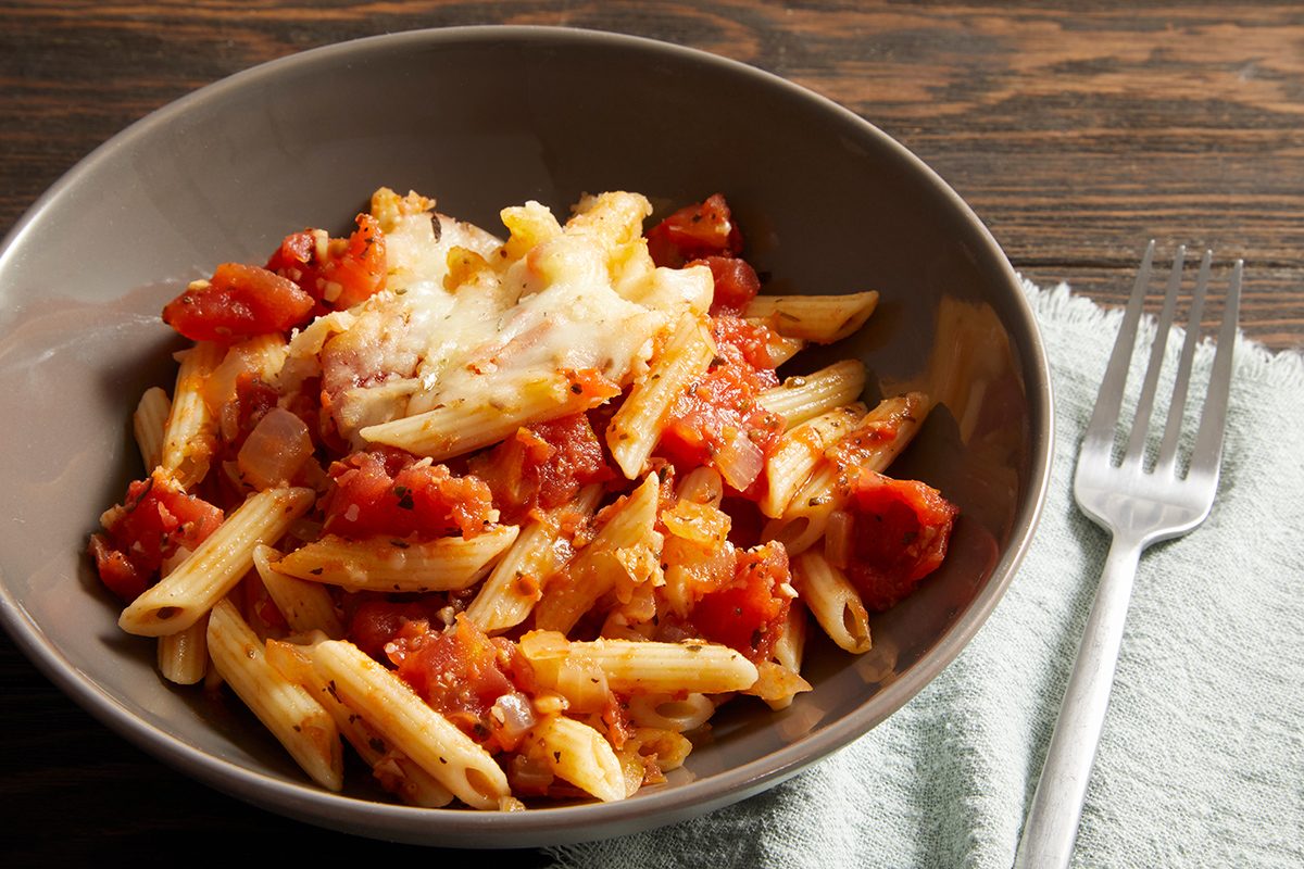 A bowl of penne pasta topped with chunky tomato sauce and melted cheese, placed on a wooden table next to a light blue napkin and a silver fork.