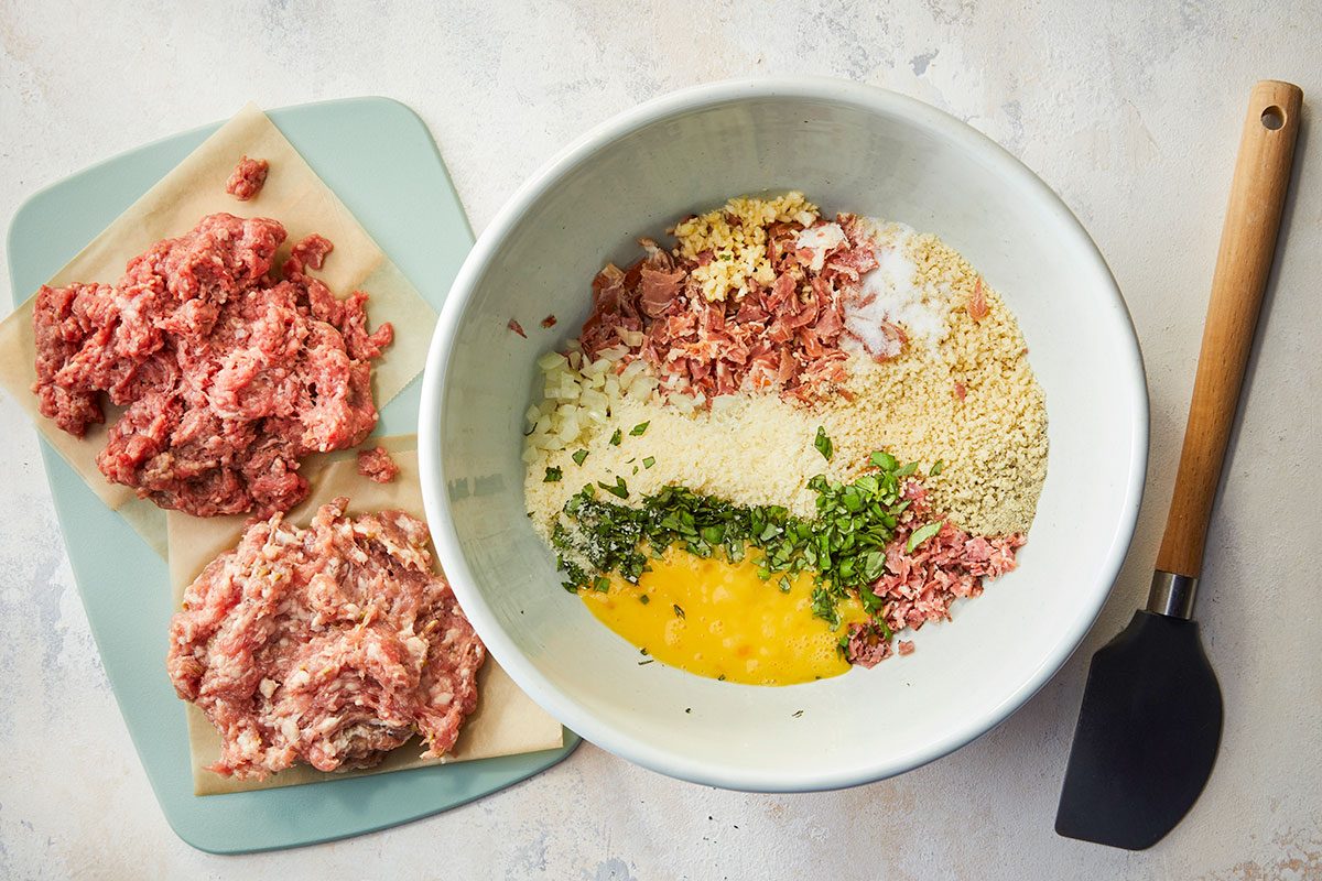 Overhead horizontal image of raw meatball mixture ingredients including ground beef, Italian sausage, eggs, herbs, breadcrumbs, and chopped cured meats in a mixing bowl.