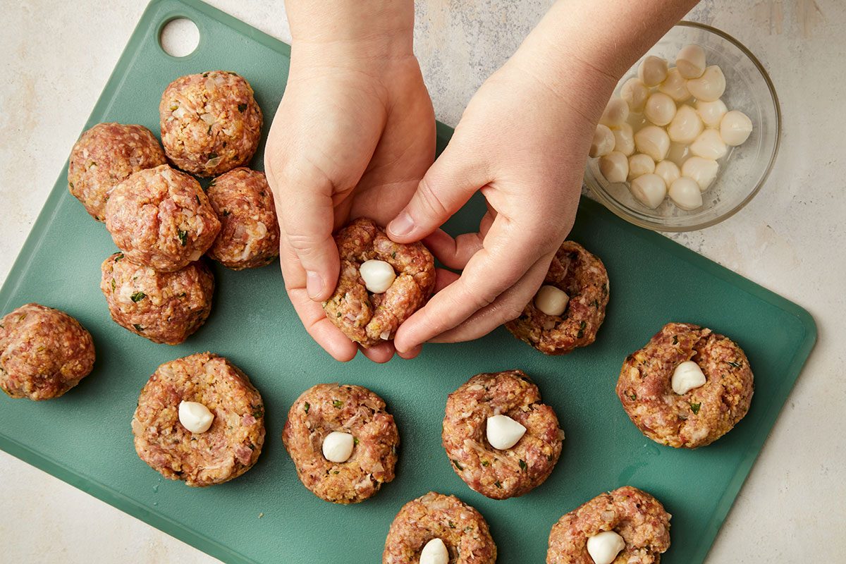 Overhead horizontal image of hands shaping meatballs and stuffing mozzarella pearls into the center of each.