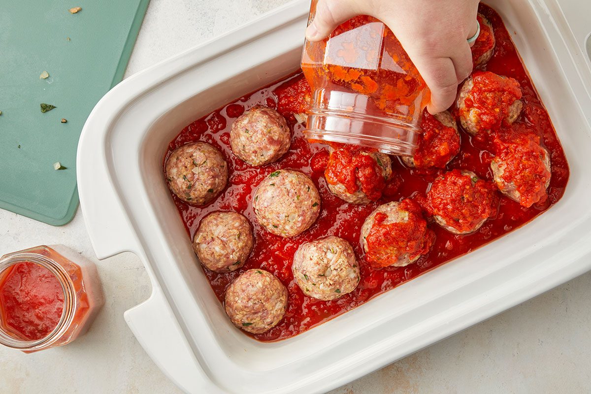 Overhead horizontal image of raw meatballs in a slow cooker covered with marinara sauce and salsa being poured over the top.