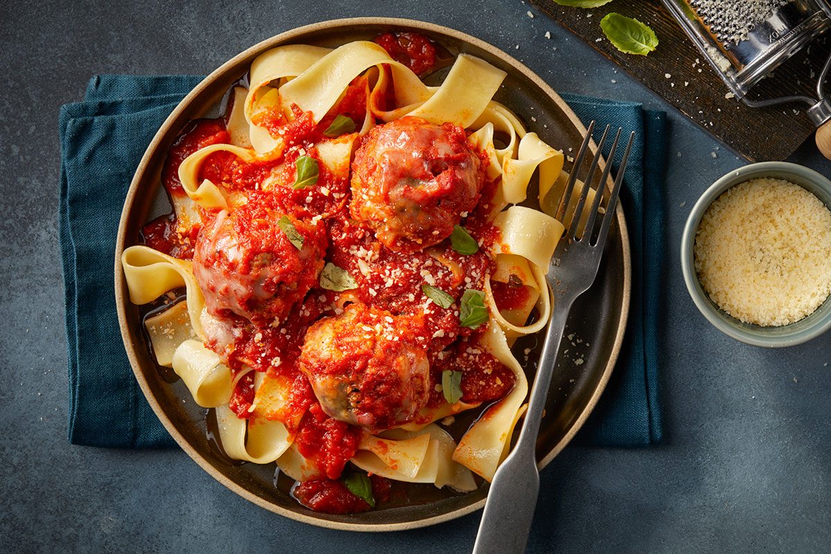 Overhead horizontal image of plated pappardelle pasta topped with multiple meatballs, marinara sauce, and grated cheese.