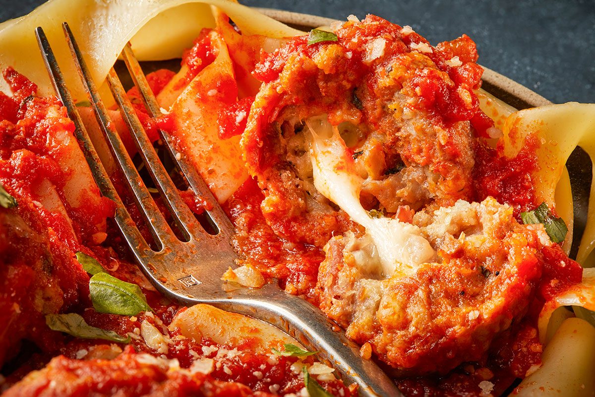 Overhead horizontal close-up of a cut meatball showing melted mozzarella center with marinara sauce and pasta.