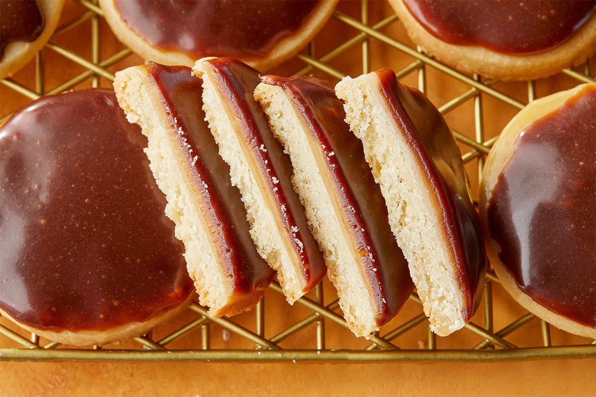 Overhead premium image featuring a sliced Twix Cookie revealing distinct layers of tender shortbread base, rich caramel filling, and smooth milk chocolate topping. The cross-section highlights texture and structure.