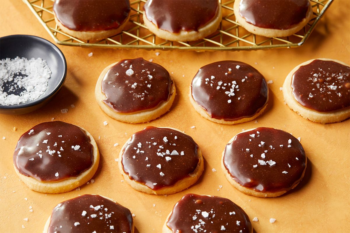 Overhead premium food photograph of Twix Cookies arranged on a warm background with a small dish of flaked sea salt. The chocolate-topped cookies are evenly coated and lightly garnished for a polished finish.