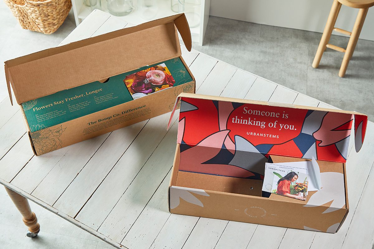 Two open cardboard boxes sit on a white wooden table. One box contains a green envelope and the other has a colorful card that says “Someone is thinking of you” along with a photo of a smiling woman.