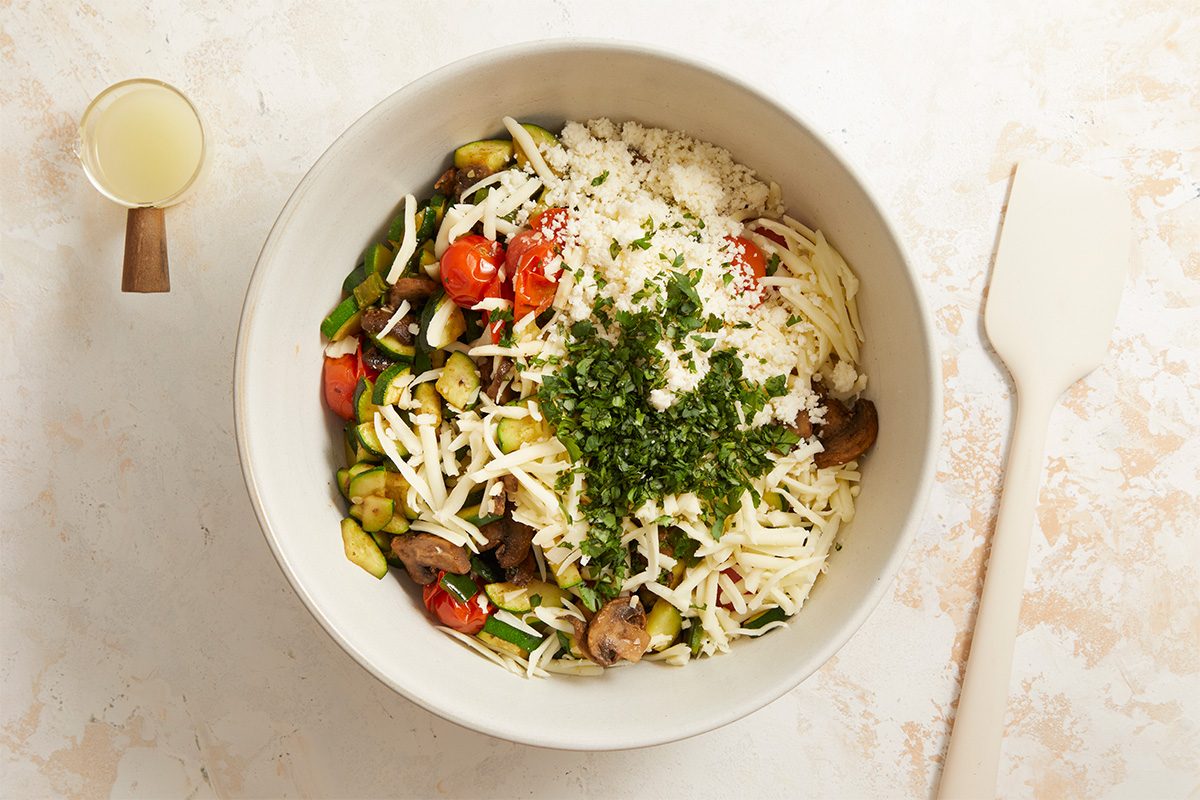 Horizontal overhead image capturing a mixed vegetable enchilada filling topped with shredded cheese and fresh herbs. This Fast Track step-by-step photograph highlights seasoning and layering during preparation of Farmers Market Enchiladas. Clean styling and bright lighting emphasize texture and ingredient clarity for instructional print and digital use.