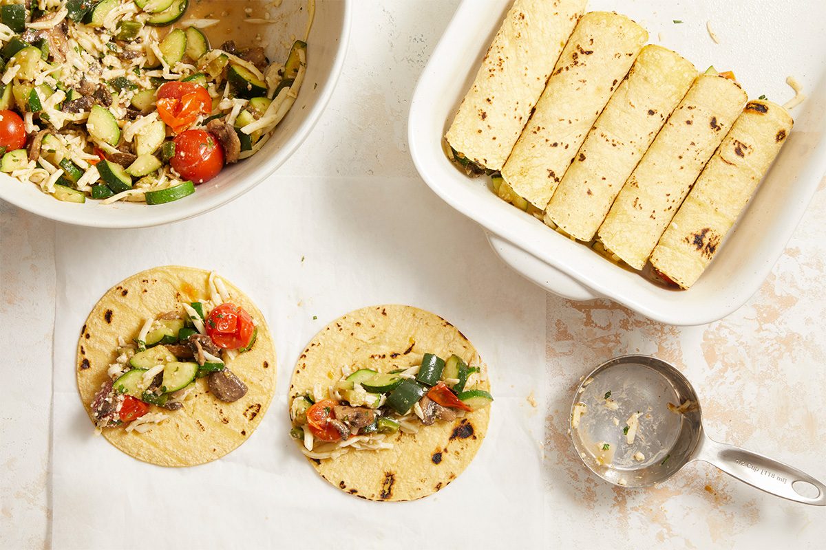 Overhead horizontal step-by-step image showing Farmers Market Enchilada filling spooned onto warmed corn tortillas. Ingredients are clearly visible, emphasizing portioning and assembly technique. This Fast Track instructional photograph supports clear recipe progression for home cooks, designed for editorial and digital recipe formats.