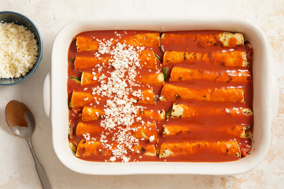 Horizontal overhead image showing rolled corn tortillas arranged in a baking dish and covered with enchilada sauce. This Fast Track step-by-step photograph highlights proper assembly before baking, emphasizing sauce coverage and structure. Designed for clear instructional storytelling in a Farmers Market Enchiladas recipe.