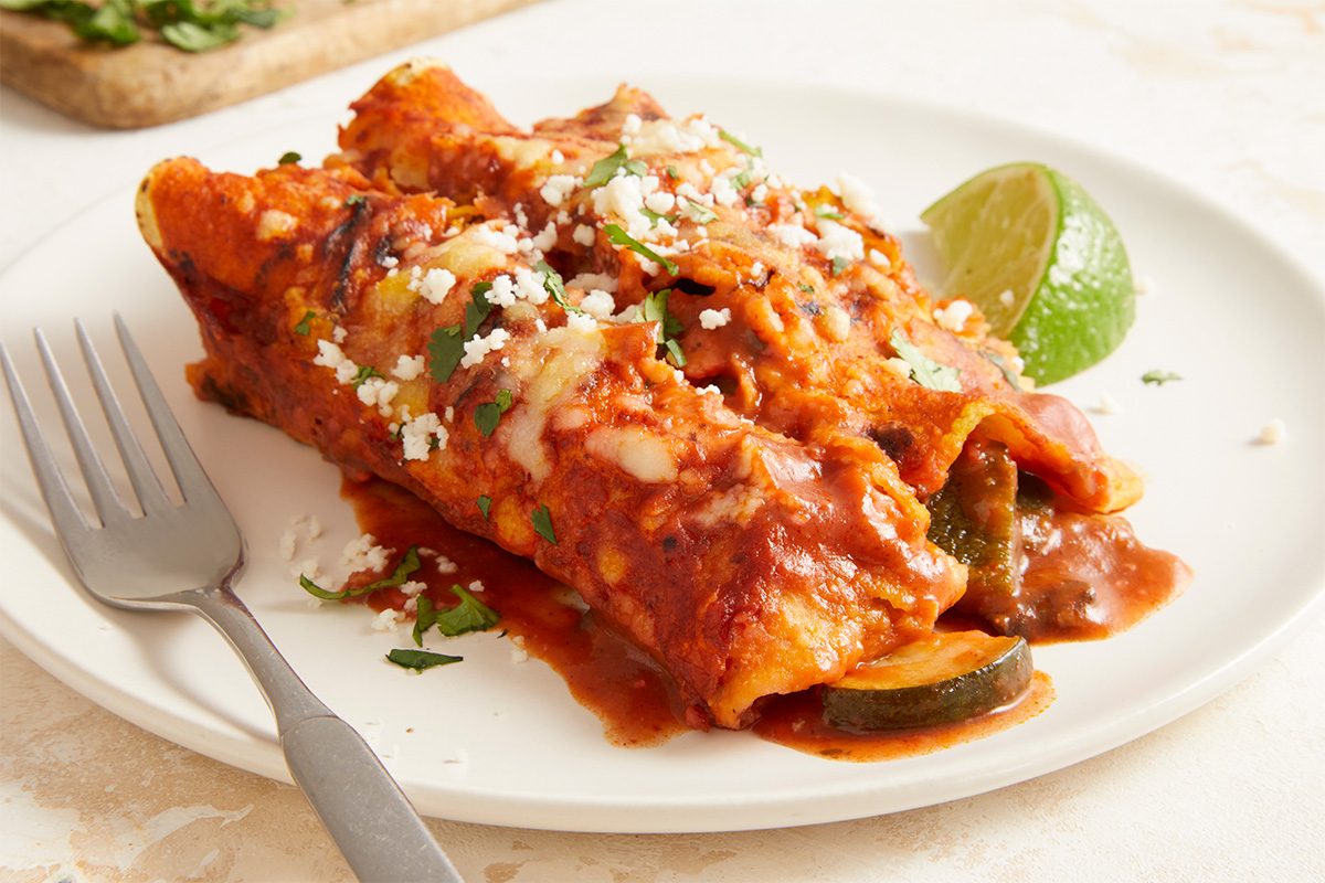 Overhead horizontal image of a plated serving of Farmers Market Enchiladas garnished with cheese and herbs. This Fast Track step-by-step photograph highlights final presentation and serving suggestion, showcasing vibrant vegetables and rich sauce for print and digital recipe publication.