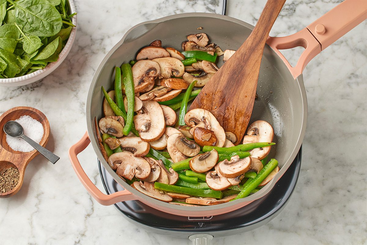 Overhead shot of sliced mushrooms and green beans sautéing in a skillet, being stirred with a wooden spatula on a stovetop set over a light marble surface;