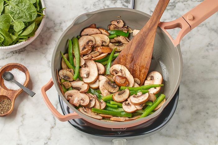 Overhead shot of sliced mushrooms and green beans sautéing in a skillet, being stirred with a wooden spatula on a stovetop set over a light marble surface;