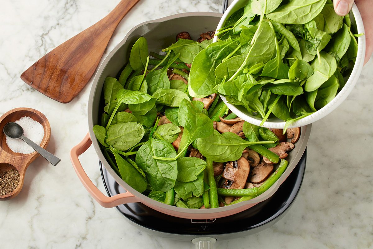 Overhead shot of fresh spinach being added to a skillet with sautéed mushrooms and green beans on a stovetop, set over a light marble surface;