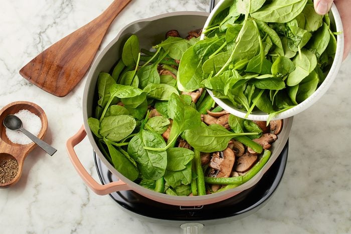 Overhead shot of fresh spinach being added to a skillet with sautéed mushrooms and green beans on a stovetop, set over a light marble surface;