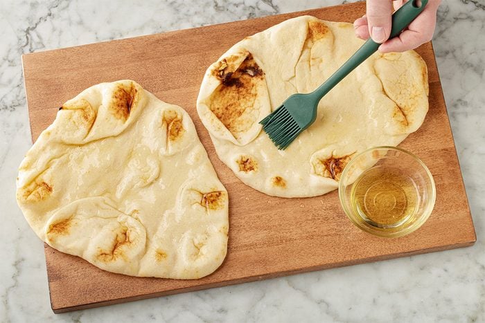 Overhead shot of flatbread being brushed with oil using a silicone brush on a wooden board, set on a light marble surface;