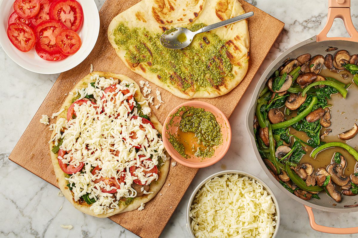 Overhead shot of flatbread pizzas in progress with pesto, shredded cheese, and sliced tomatoes on a wooden board, alongside a skillet of sautéed mushrooms and greens on a light marble surface;