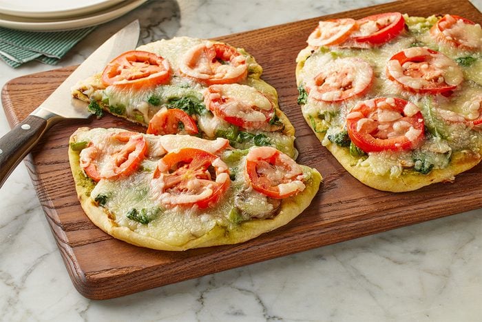 3/4 angle shot of grilled flatbread veggie pizzas topped with melted cheese, sliced tomatoes, and pesto, served on a wooden board over a light marble surface;