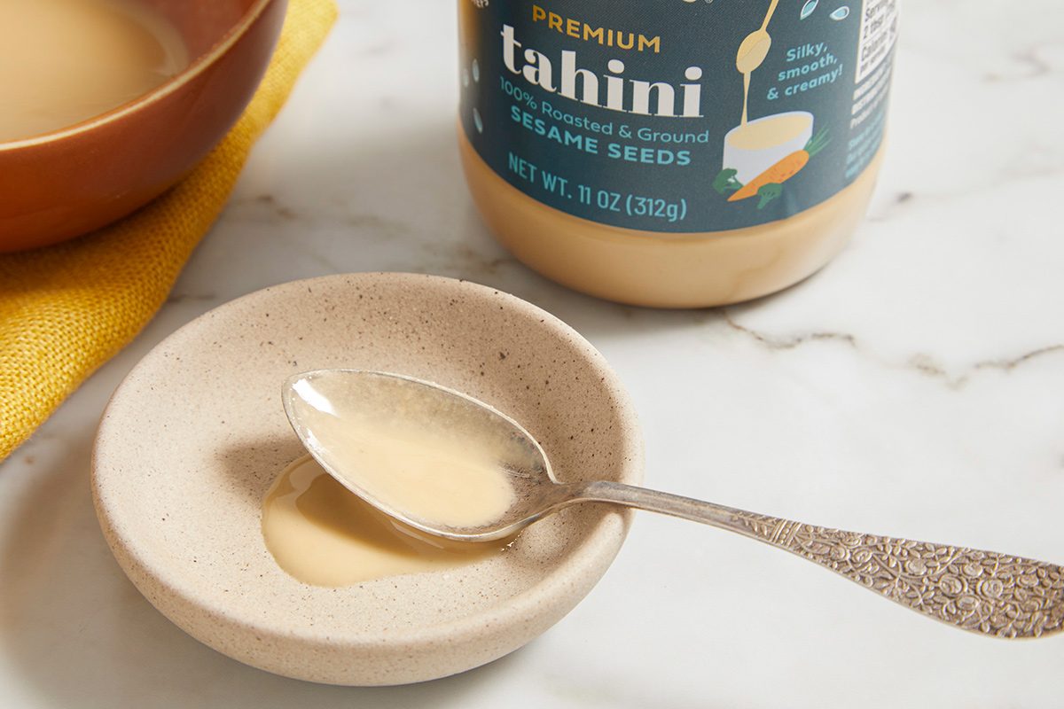A spoon with creamy tahini rests on a small ceramic dish next to an open jar of tahini on a marble countertop. A yellow cloth and a brown bowl are partially visible in the background.