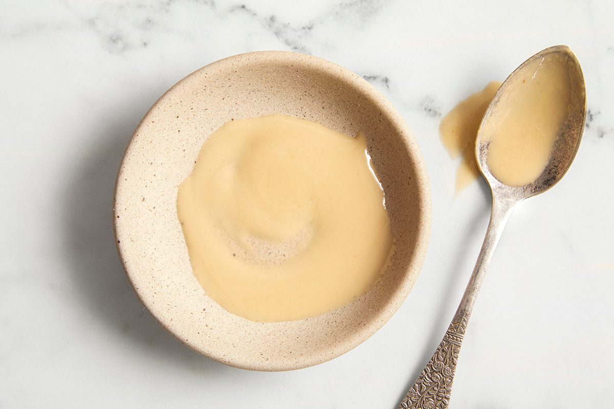 A small beige bowl filled with creamy beige tahini sits on a white marble surface, with a silver spoon holding some tahini placed beside it.