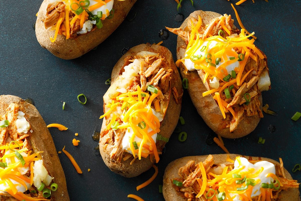 Baked potatoes topped with shredded chicken, sour cream, shredded cheddar cheese, and chopped green onions on a dark surface.